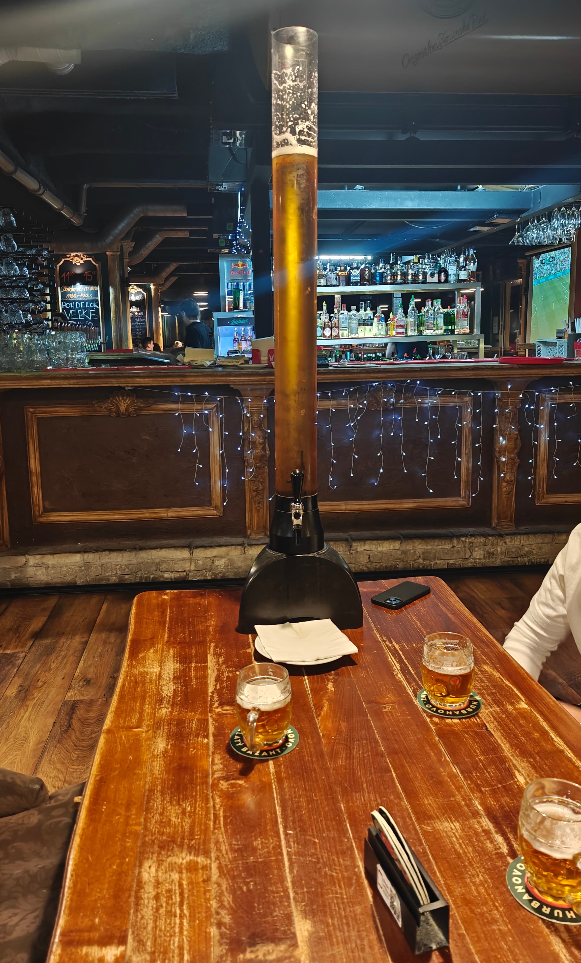 A tall self-pour beer tower on a pub table, with small glasses on coasters and the bar behind