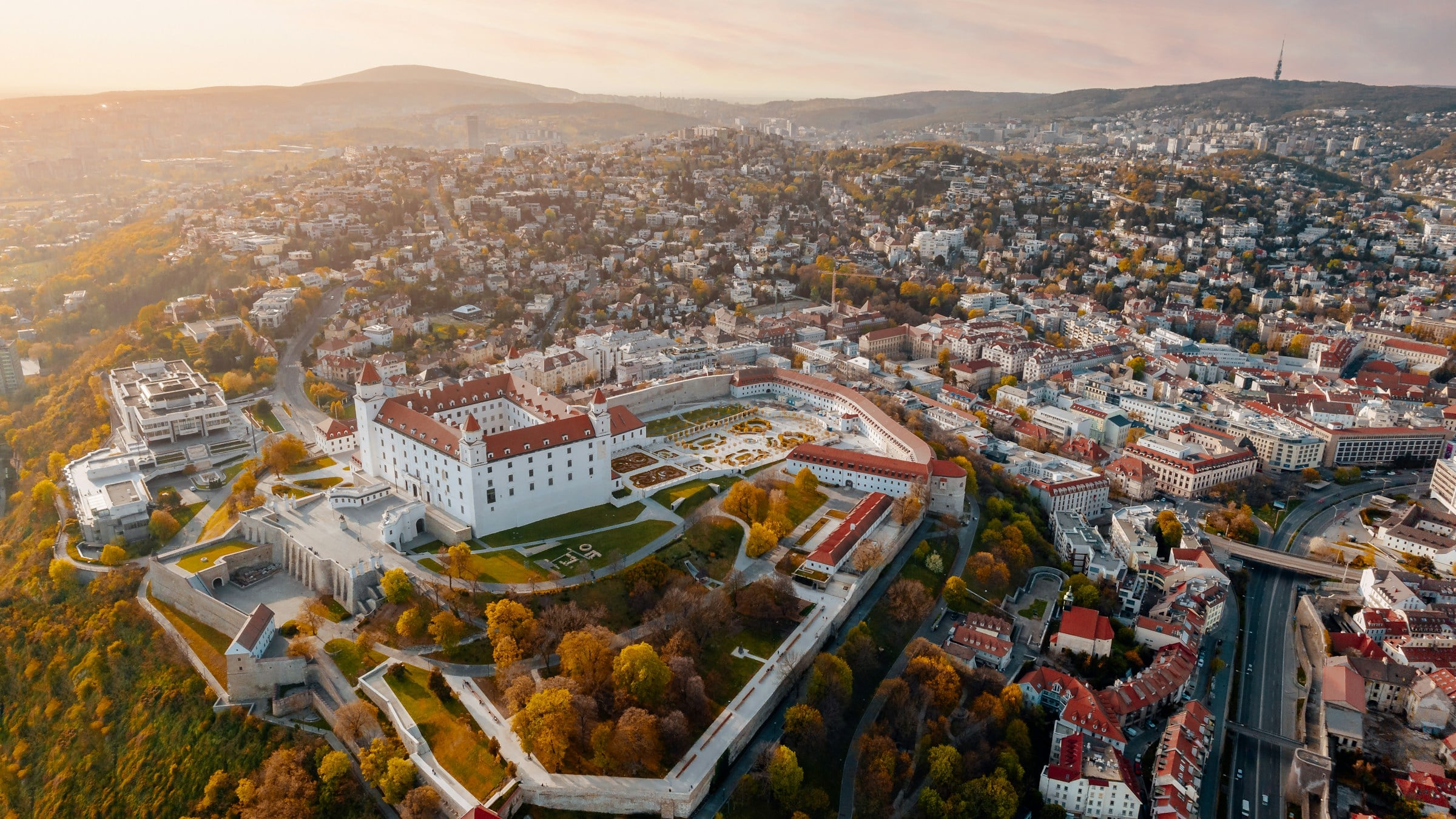 Aerial view of Bratislava at sunset: the white castle with its red roofs and baroque garden, the Old Town rooftops beside it, wooded hills on the horizon
