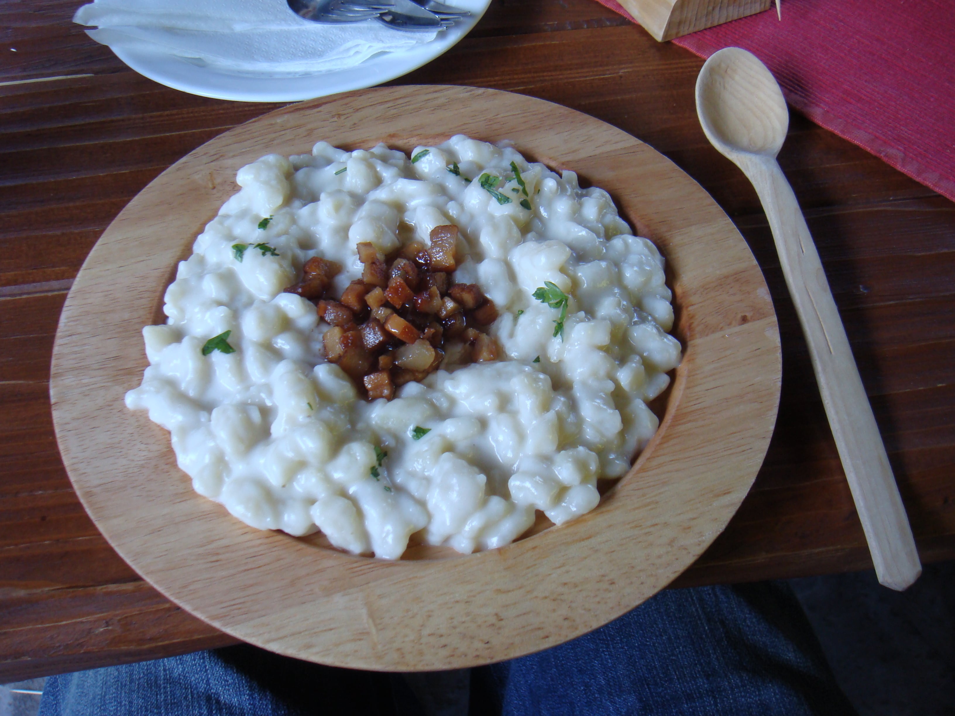 A wooden bowl of bryndzové halušky topped with fried bacon cubes and chopped herbs, a wooden spoon beside it