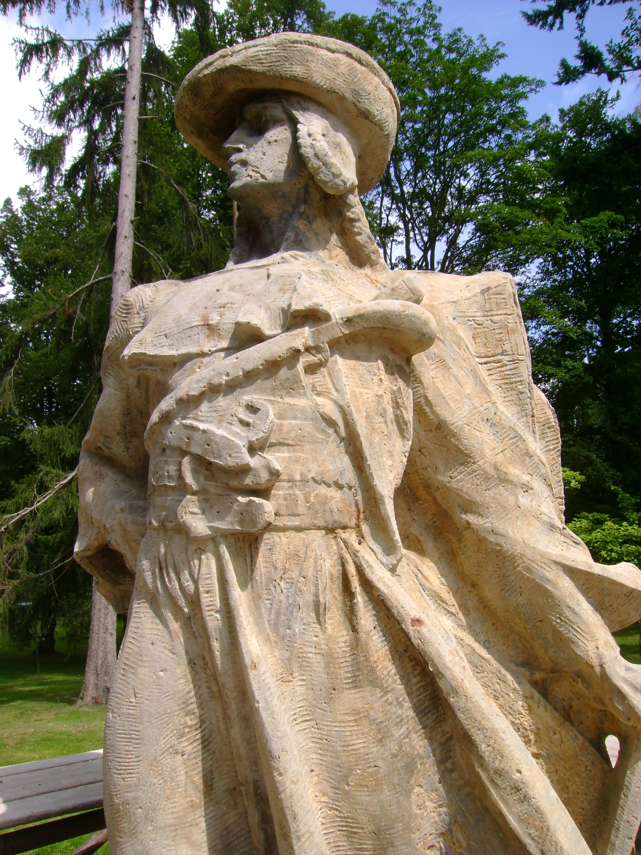 Stone statue of Jánošík in a wide-brimmed shepherd hat and long coat, seen from below against trees