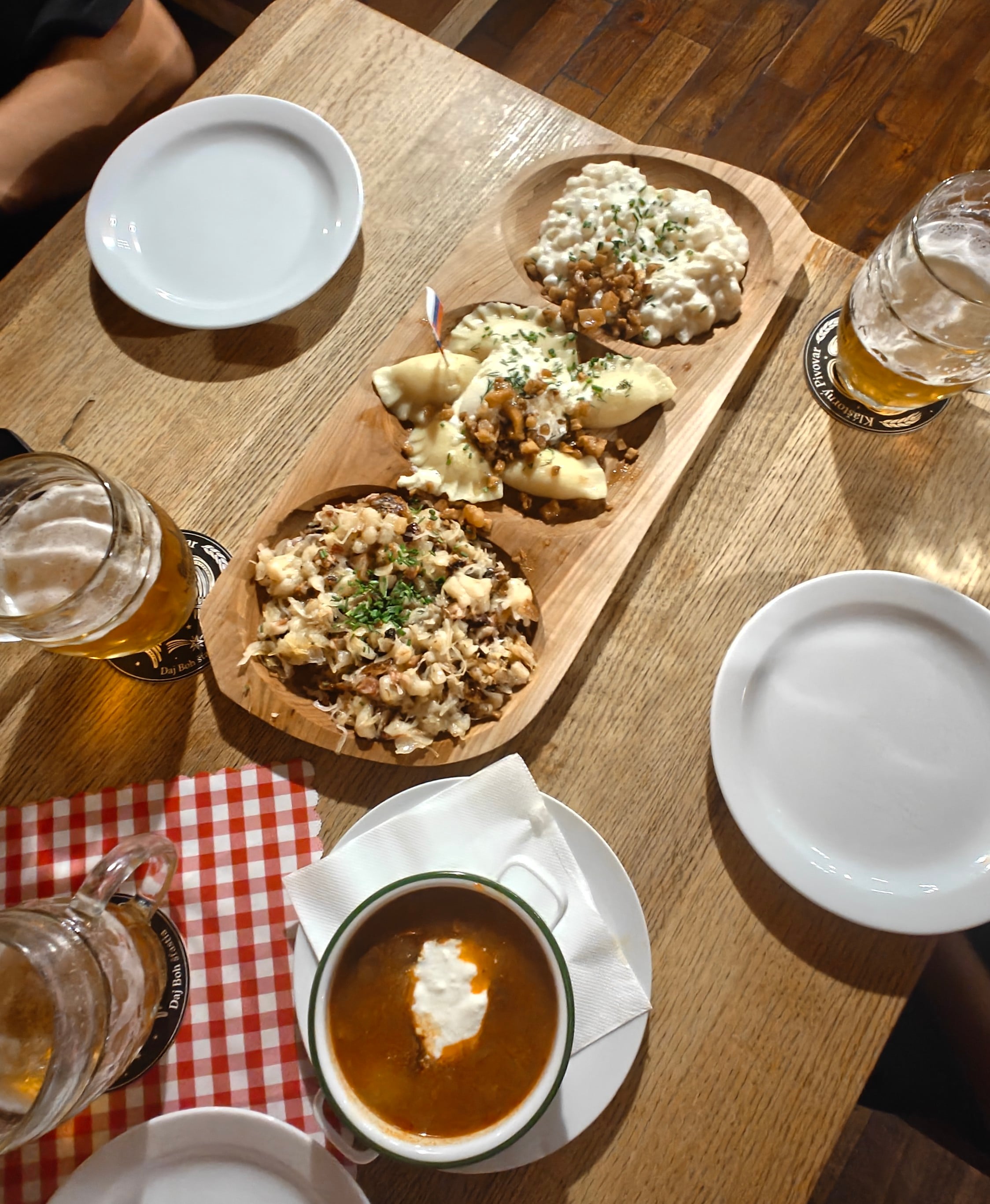 Wooden sharing board with bryndzové halušky, pirohy and strapačky, next to a cup of kapustnica and glasses of beer