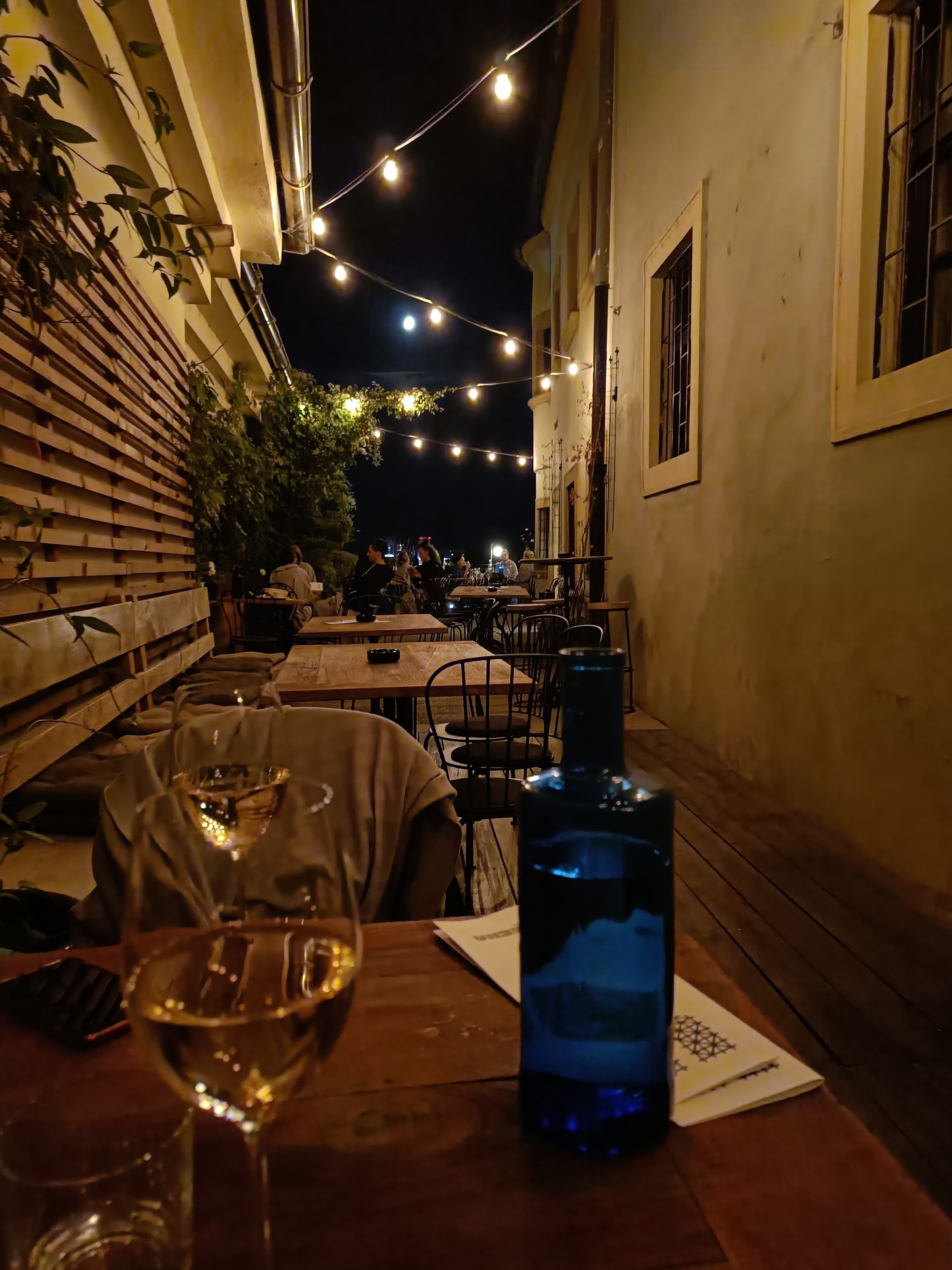 Narrow terrace at Viecha pod hradom at night, lit by string lights, with a glass of white wine in the foreground