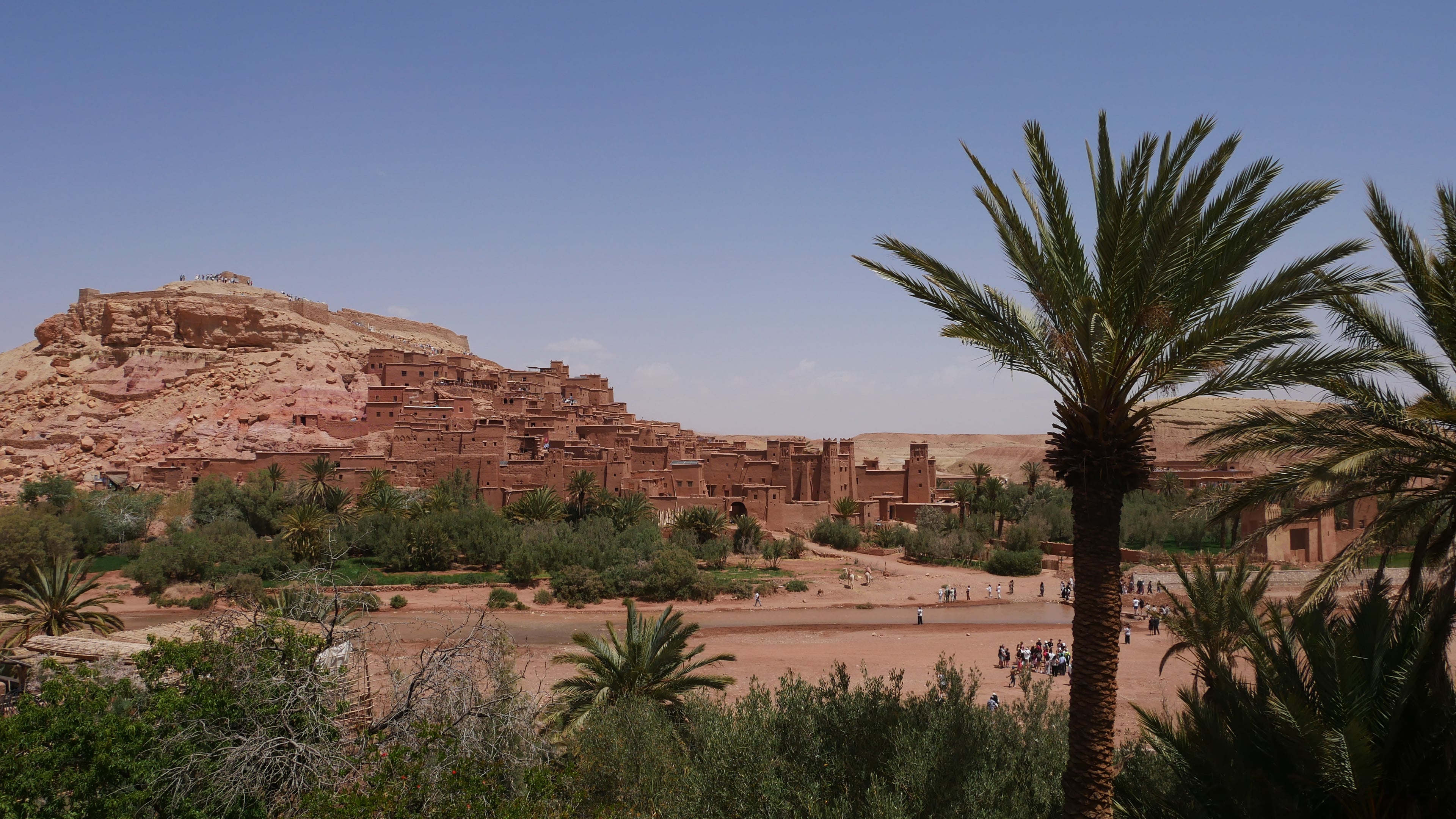 The fortified mud-brick town of Ait Ben Haddou rising up a hillside above palm trees and a dry riverbed