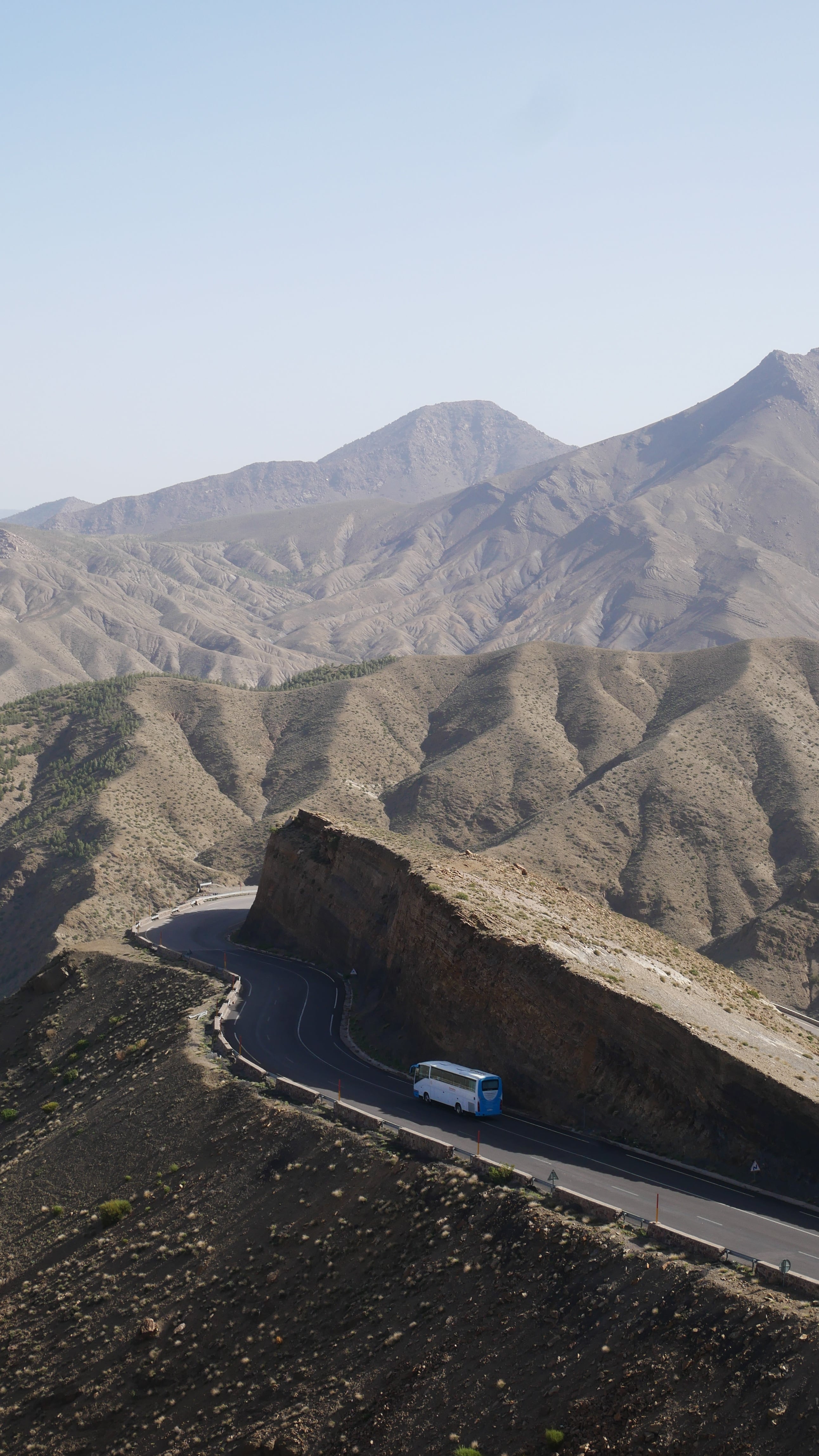 A coach on a hairpin bend of a mountain road cut into a bare ridge of the High Atlas, with layered peaks behind
