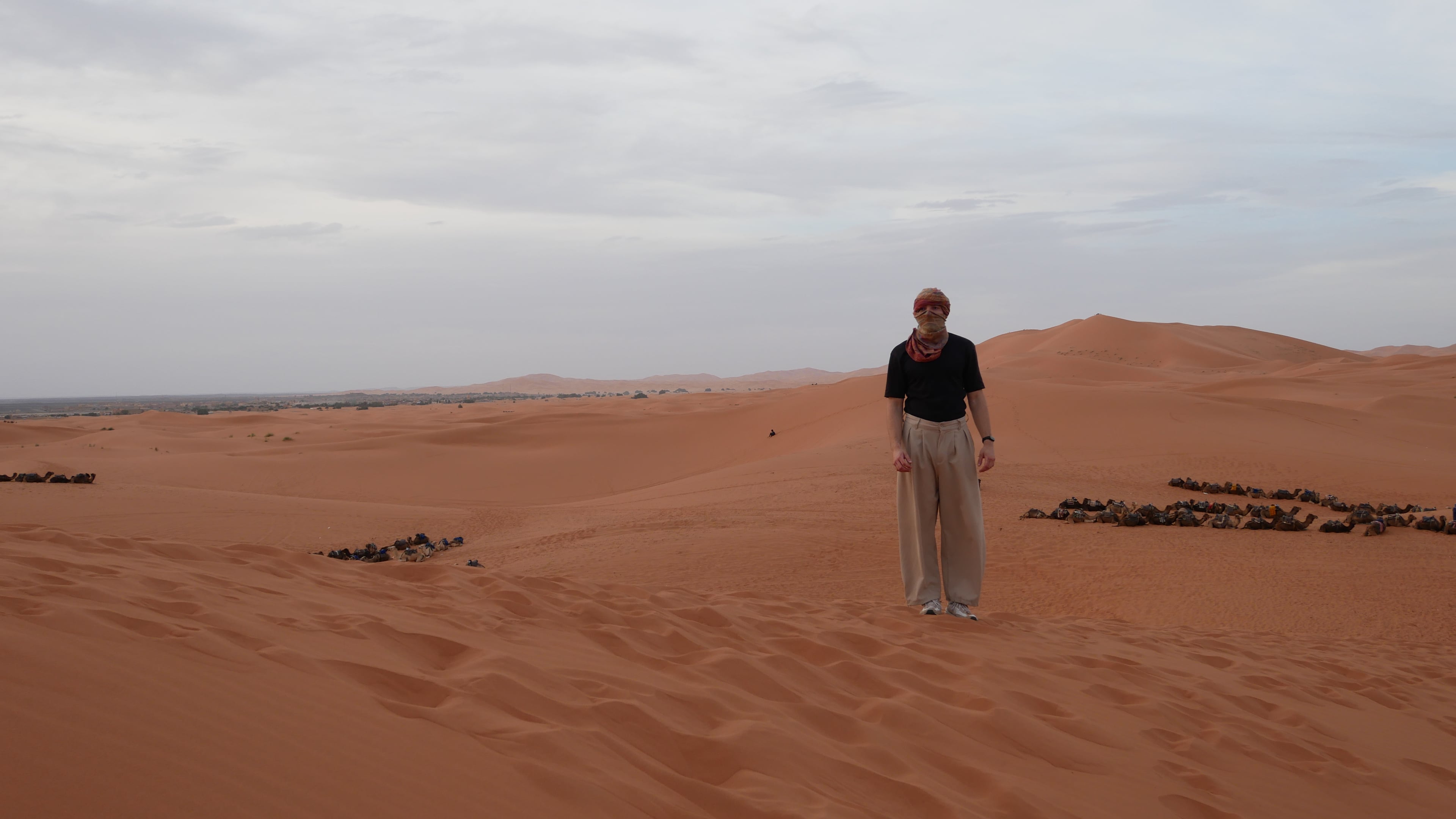 The author standing on a sand dune in a turban, with camels resting on the sand behind him and a village visible on the horizon