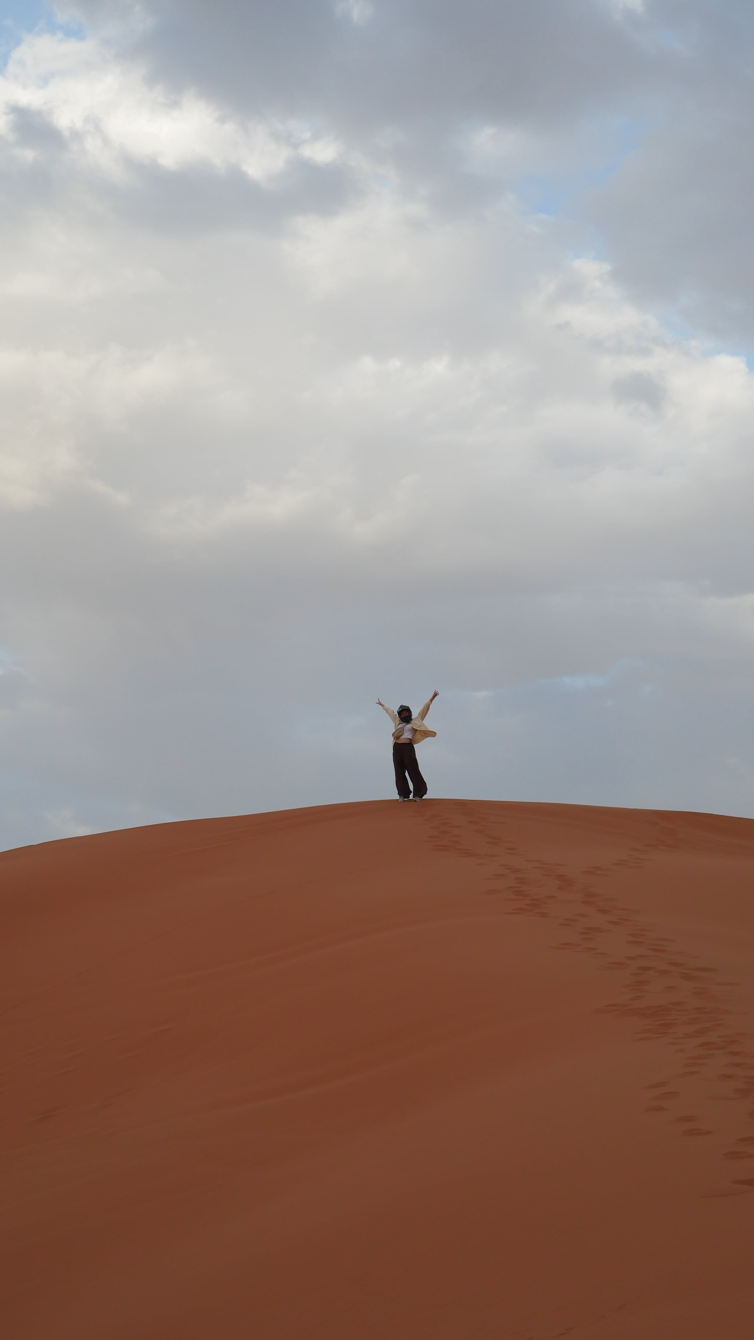 A person standing with arms raised on the crest of a large red sand dune, their footprints leading up the slope behind them