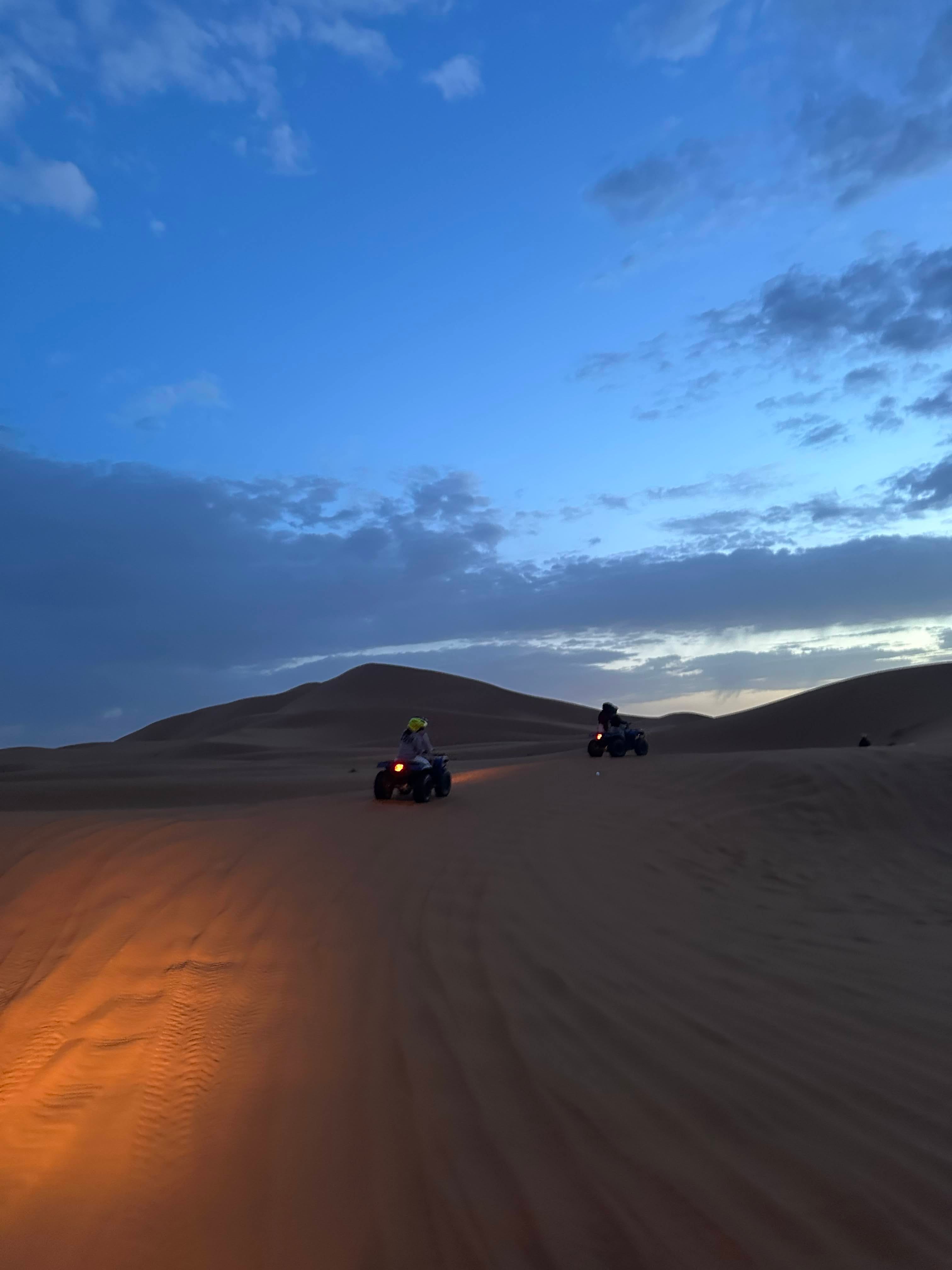 Two people on quad bikes riding across the dunes at dawn, headlights on, under a deep blue sky streaked with cloud