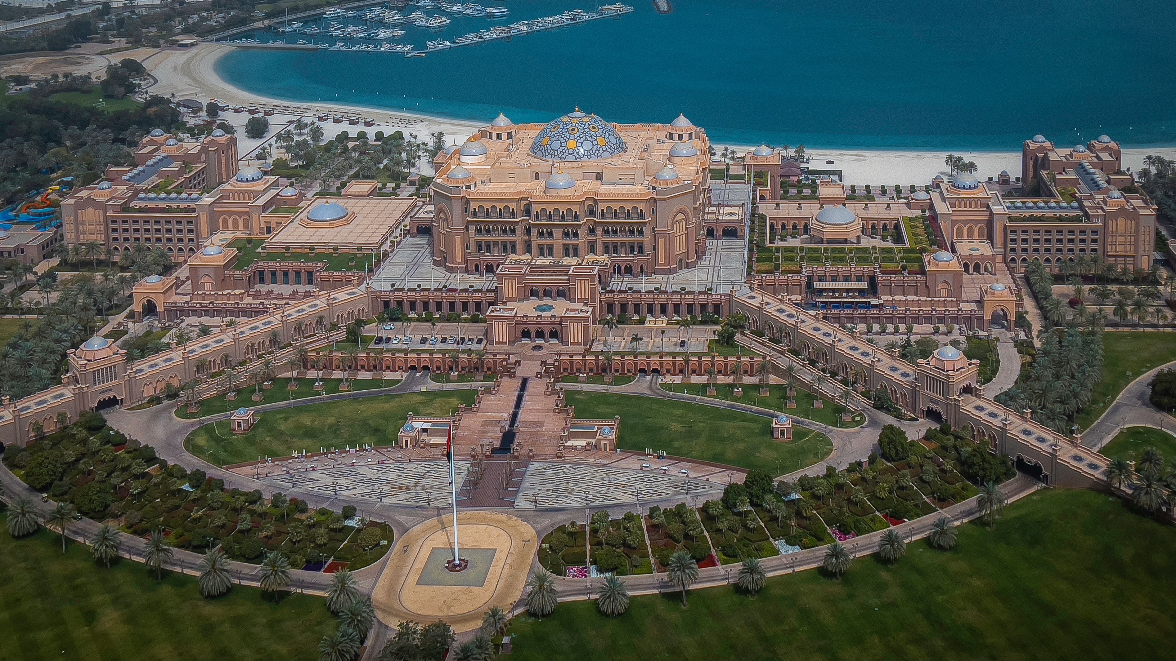 Aerial view of the Emirates Palace: a long symmetrical sandstone palace with domes and formal gardens, facing the Arabian Gulf