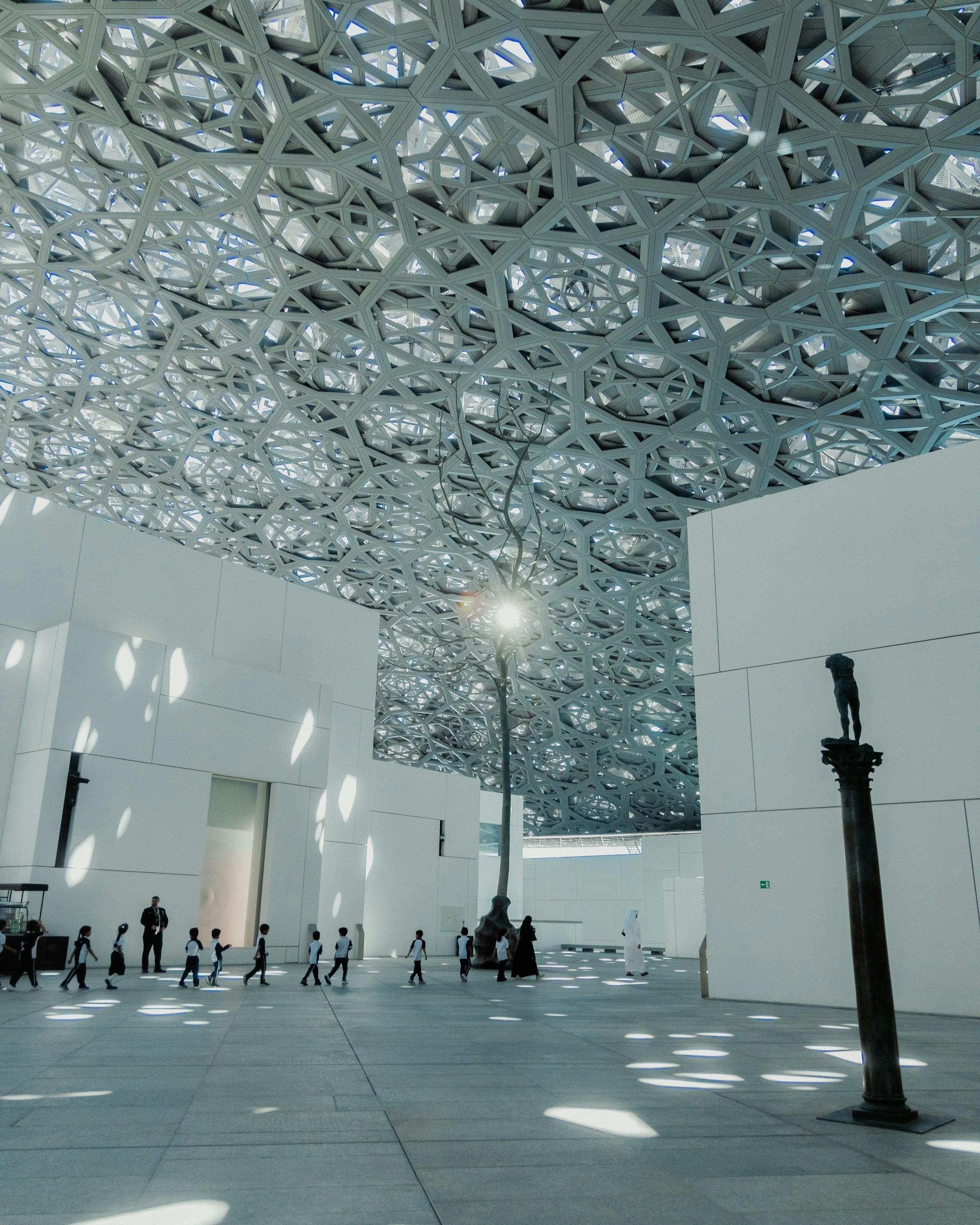 Visitors walking under the perforated metal dome of the Louvre Abu Dhabi, which scatters hundreds of spots of sunlight across the white walls and floor