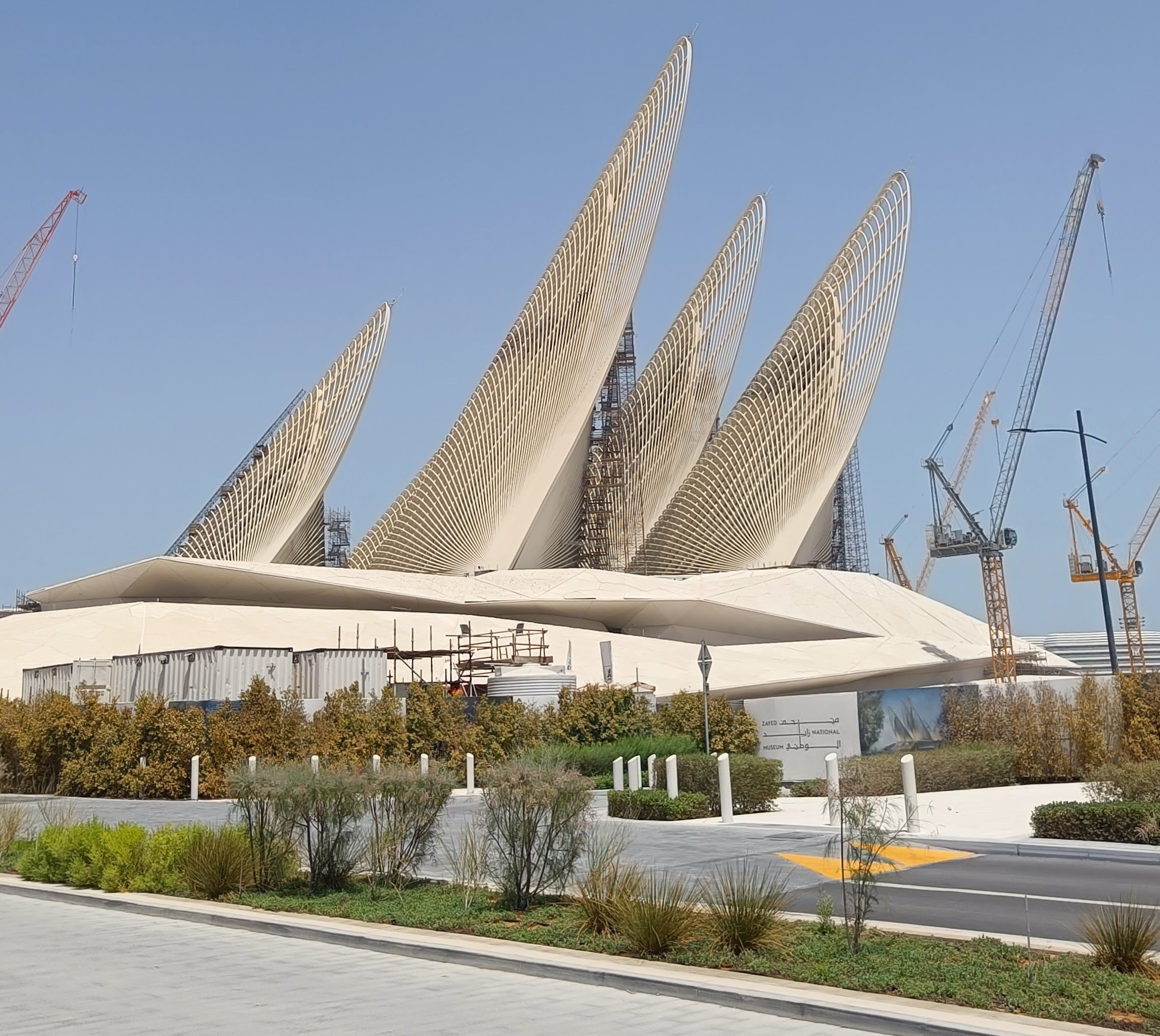 Five tall steel feather-shaped towers rising from the low white roof of the Zayed National Museum, surrounded by construction cranes